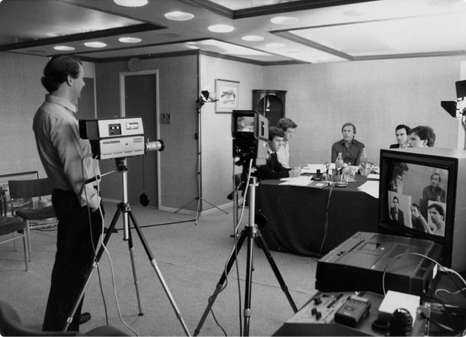 Black-and-white vintage scene of a filmed meeting, with a cameraman recording four men seated at a table, surrounded by studio lights, tripods, and video equipment in an office setting